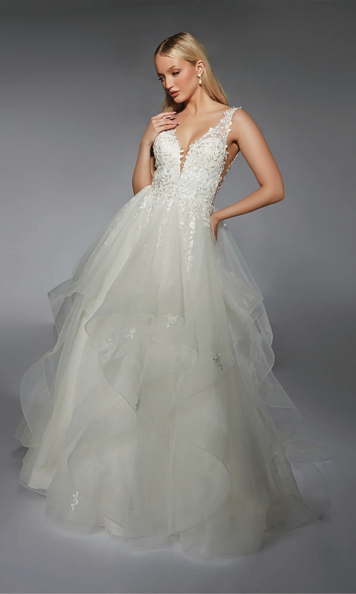 A woman in the Alyce 7126 long white dress with a deep V-neckline, floral lace details, and a layered tulle skirt stands against a gray background.
